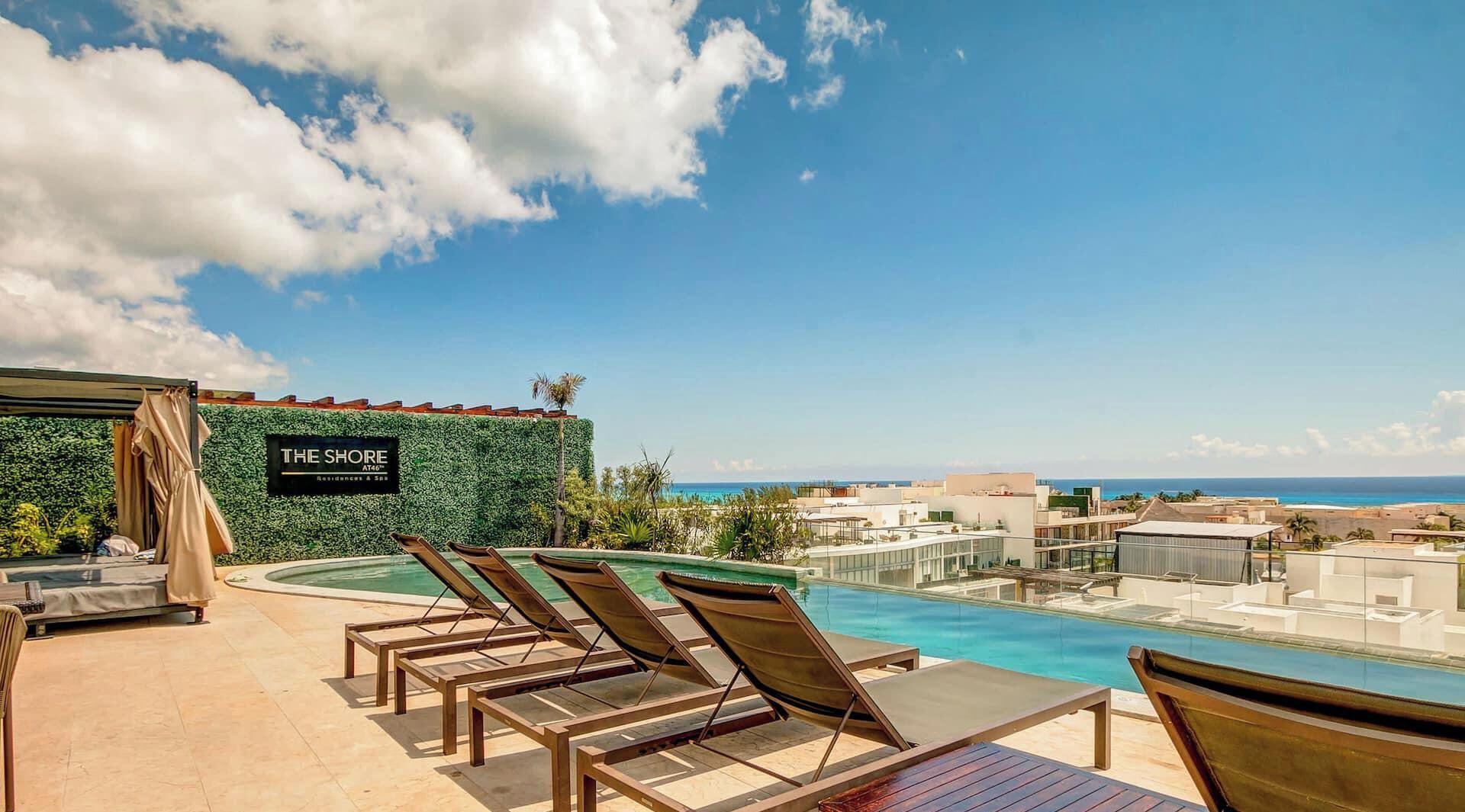 A rooftop pool area with lounge chairs, a sign reading 'THE SHORE Residences & Spa', and a view of the ocean and cityscape.