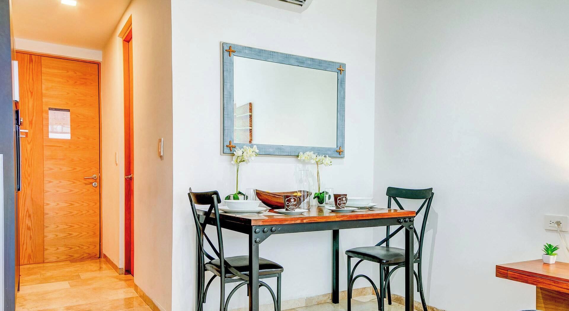 A dining area with a wooden table, two chairs, a mirror, and a small potted plant.