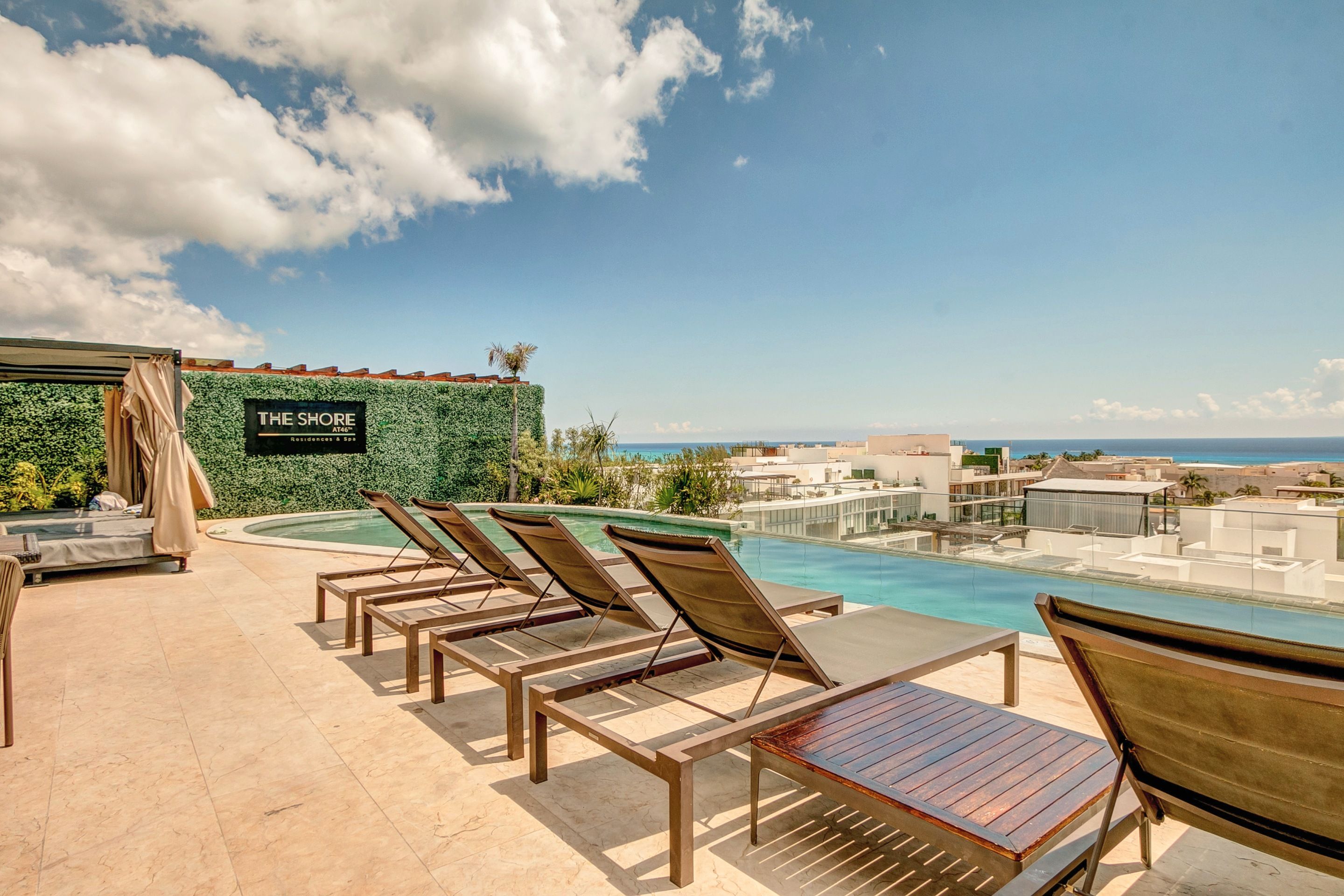 A rooftop pool area with lounge chairs, a sign for 'The Shore Residences & Spa', and a view of the ocean.
