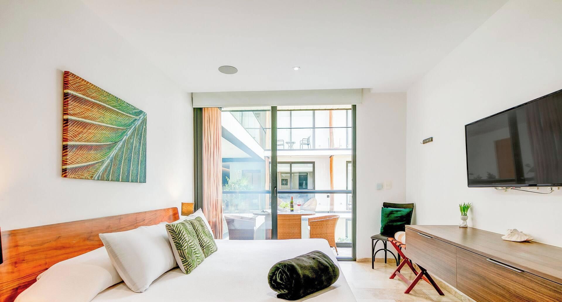 A hotel room with a bed, a TV, a desk, and a view through a glass door.