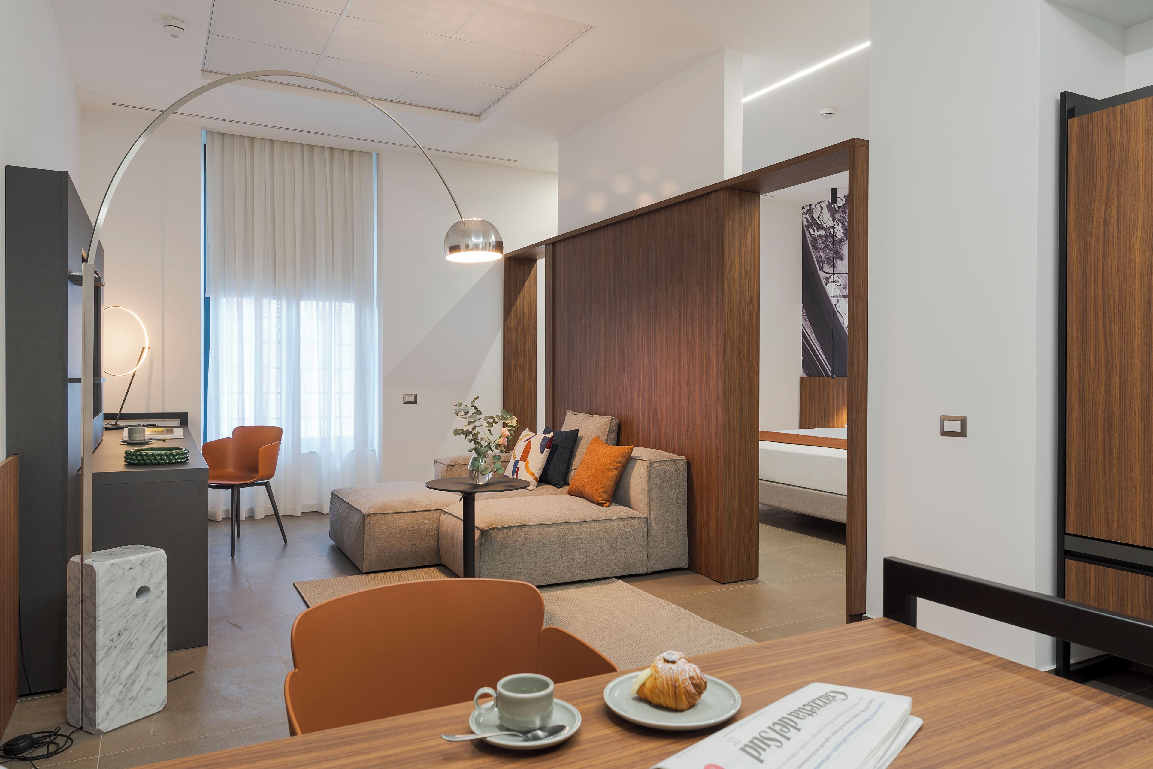 Ett modernt hotellrum med en soffa, ett matbord med en kopp och ett bakverk, ett skrivbord med en stol och ett stort fönster med gardiner.