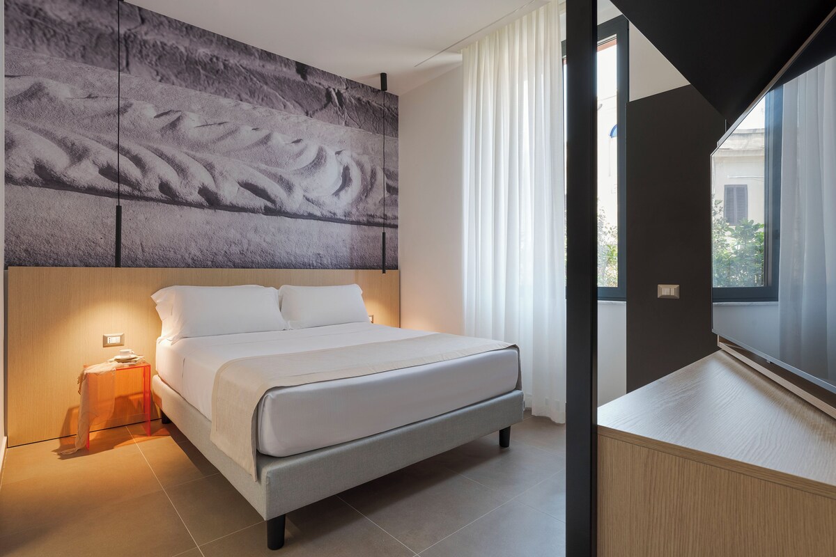Quarto moderno com cama, mesa de cabeceira, obra de arte na parede e vista para o exterior pela janela.