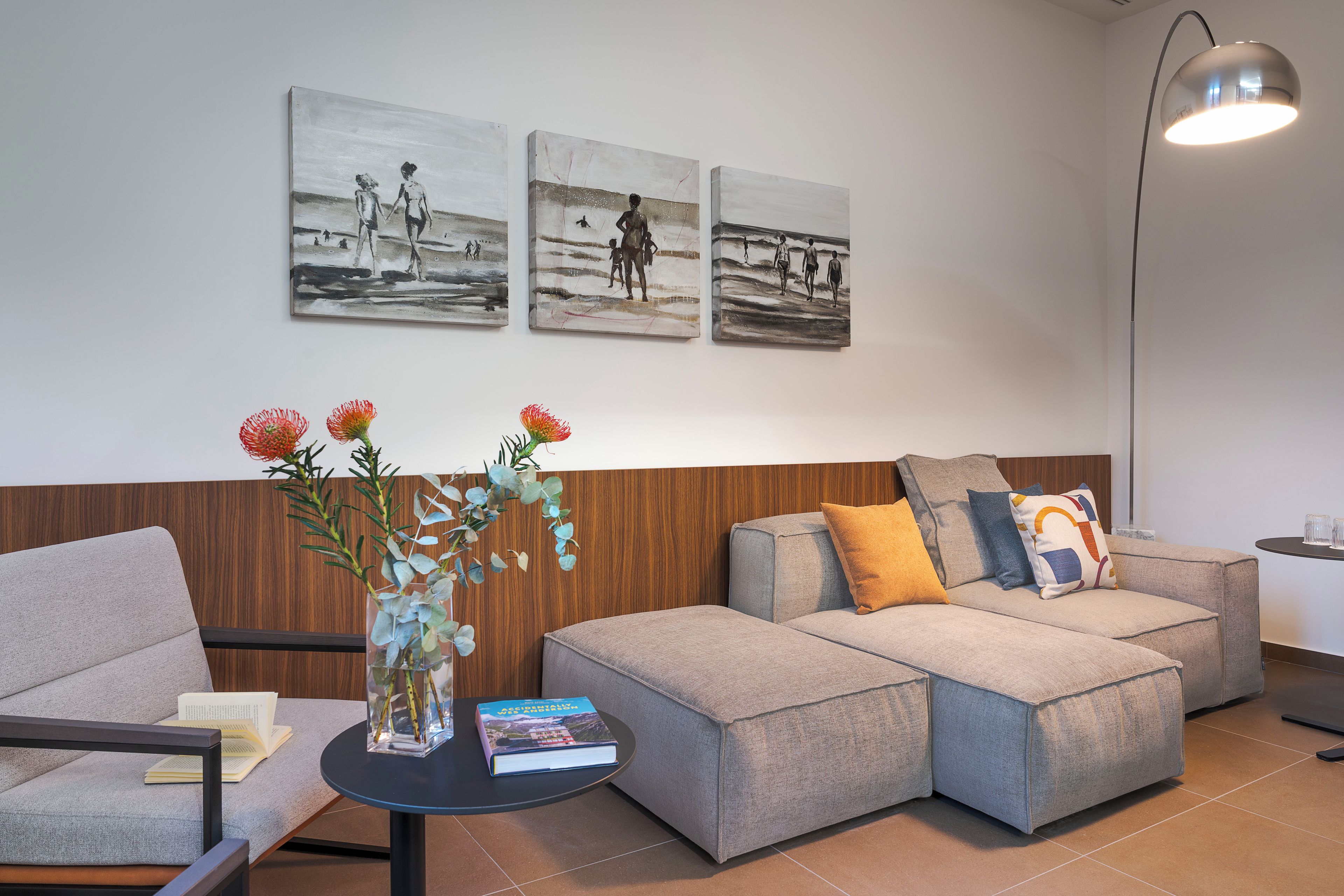 Un salon moderne avec un canapé d’angle gris, une petite table d’appoint sur laquelle se trouvent un livre et un vase de fleurs, et trois tableaux en noir et blanc accrochés au mur.