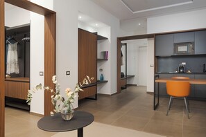 Quarto de hotel moderno com guarda-roupa de madeira, uma pequena mesa redonda com um vaso de flores, área de cozinha com micro-ondas e uma cadeira laranja.