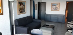 Ein modernes Wohnzimmer mit einer Couch, einem kleinen Tisch und einem Sessel. Es gibt eine Küchenzeile mit Schränken und einer Arbeitsplatte. Zwei Bilder hängen an der Wand.