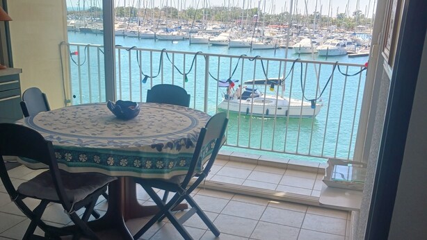 Un espace repas comprenant une table et des chaises, une vue sur une marina avec des bateaux, et un sol carrelé.