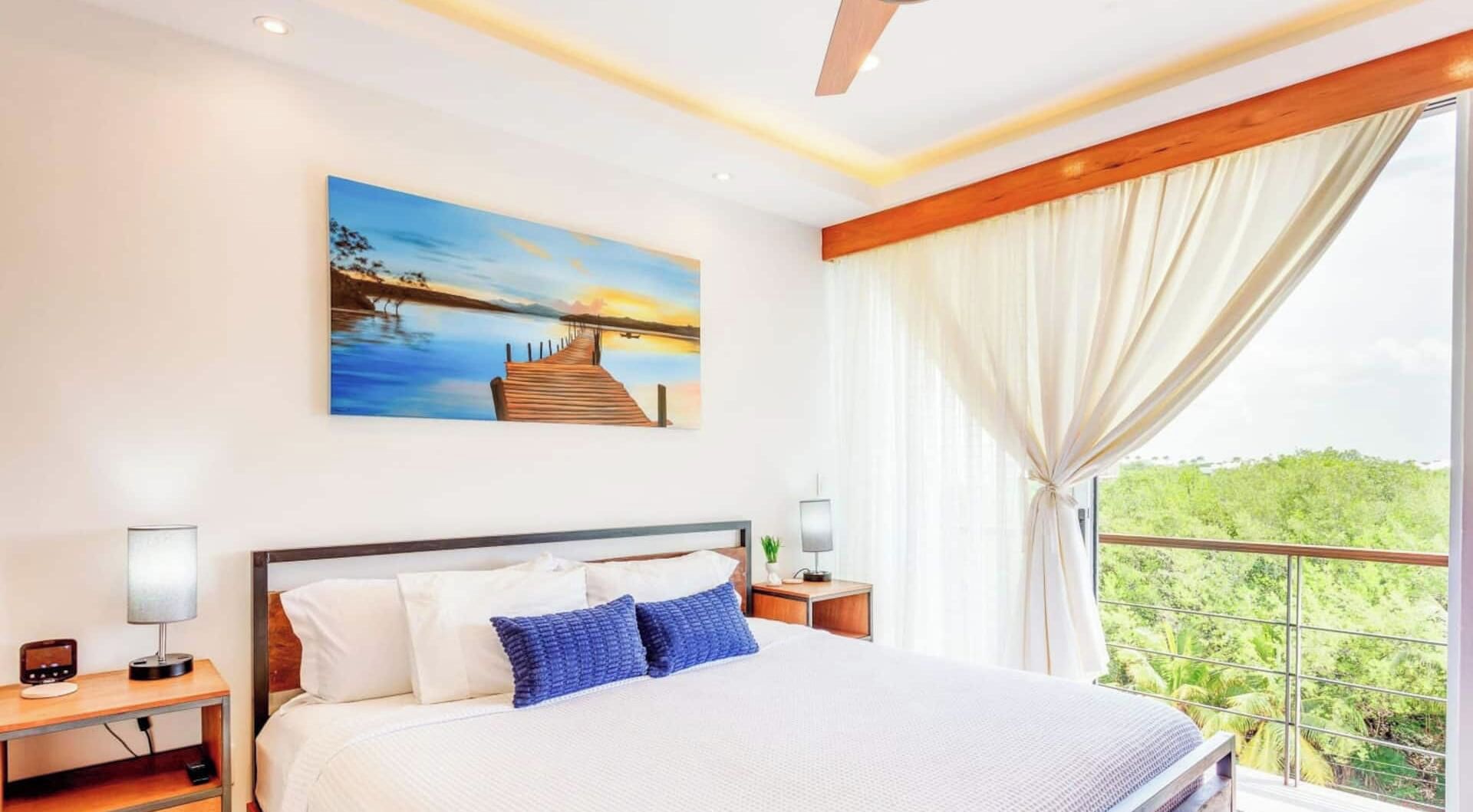 Hotelkamer met een bed, een schilderij van een zonsondergang boven het water, een nachtkastje met een lamp en uitzicht op groen door een raam.