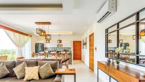 A modern living room with a sofa, dining area, kitchen, and a large window with curtains. - Beachside retreat w/ rooftop pool & BBQ (Playa del Carmen)