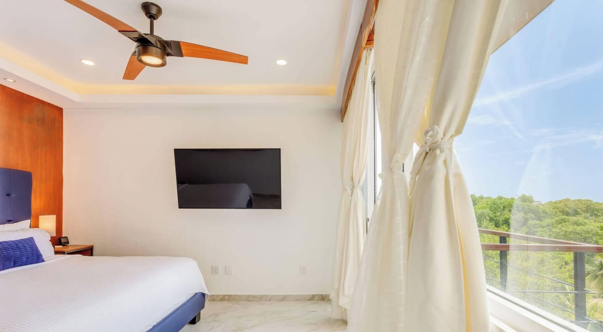 Ein Schlafzimmer mit einem Bett, einem Deckenventilator, einem Wand-Fernseher und einem Balkon mit Vorhängen.