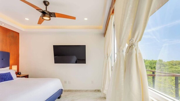 A bedroom with a bed, a ceiling fan, a mounted TV, and a balcony with curtains. - Beachside retreat w/ rooftop pool & BBQ (Playa del Carmen)