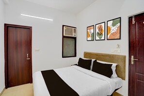 Ein Hotelzimmer mit einem Bett, einer Holztür, einer Klimaanlage und drei gerahmten Kunstwerken an der Wand.