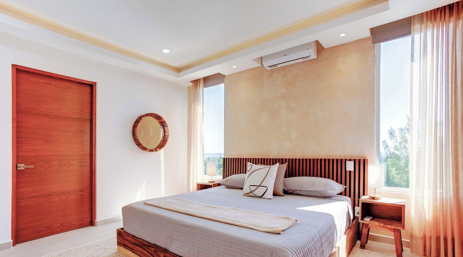 A bedroom with a large bed, a wooden headboard, a wall-mounted air conditioning unit, and a view of the outdoors through a window.