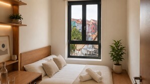 A bedroom with a bed, bedside table, chair, window, and a view of buildings outside. - Maison Bruxelles (Schaerbeek)