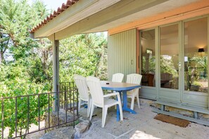 Un espace extérieur abrité, aménagé avec une table et des chaises, une rambarde en métal, et offrant une vue sur des arbres et un bâtiment en arrière-plan.