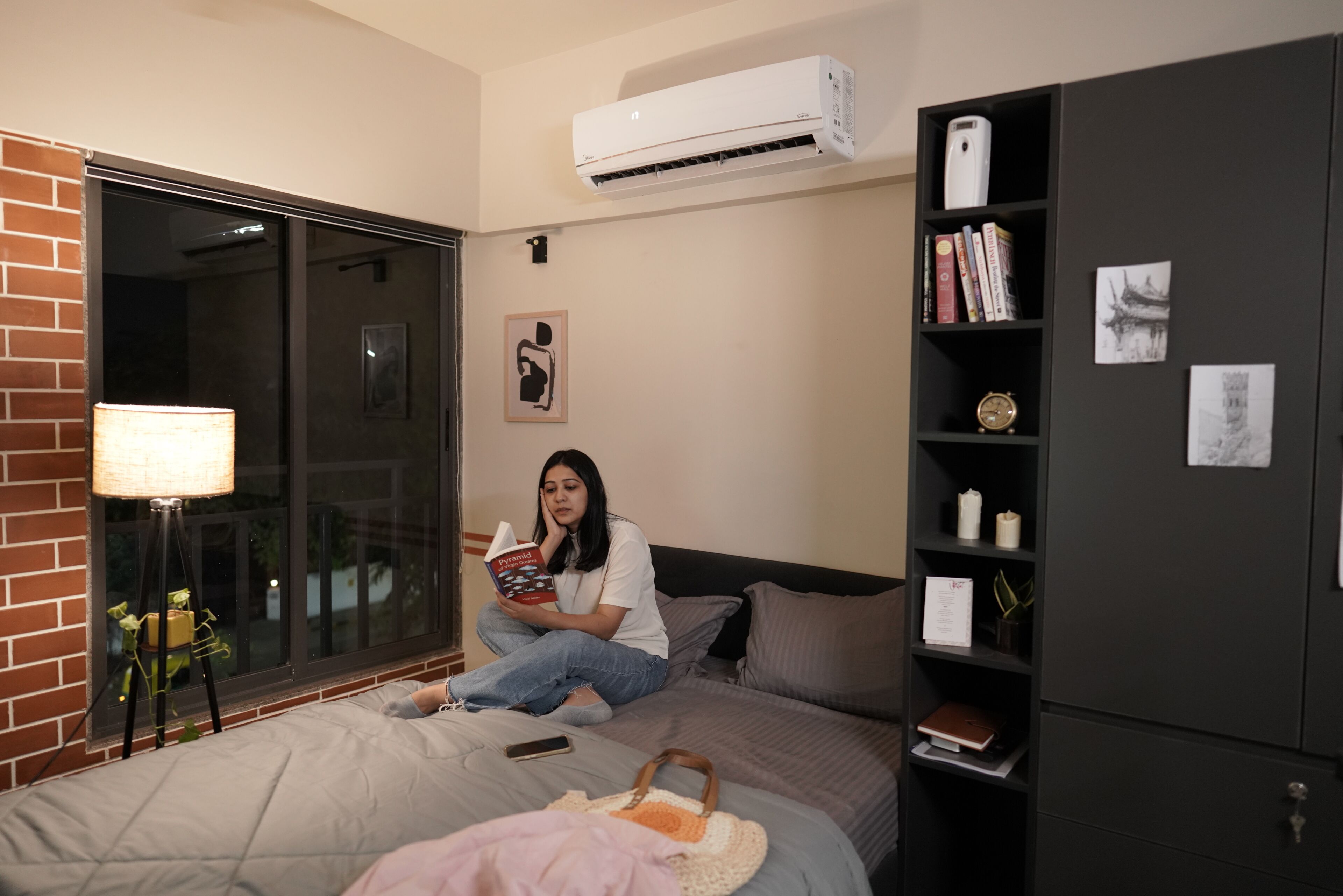 A room with a bed, a bookshelf, a window with a view, and an air conditioner.