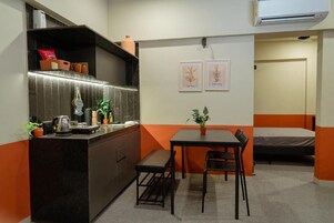 A compact room with a kitchenette, a dining table with chairs, a bed, and two framed botanical prints on the wall.
