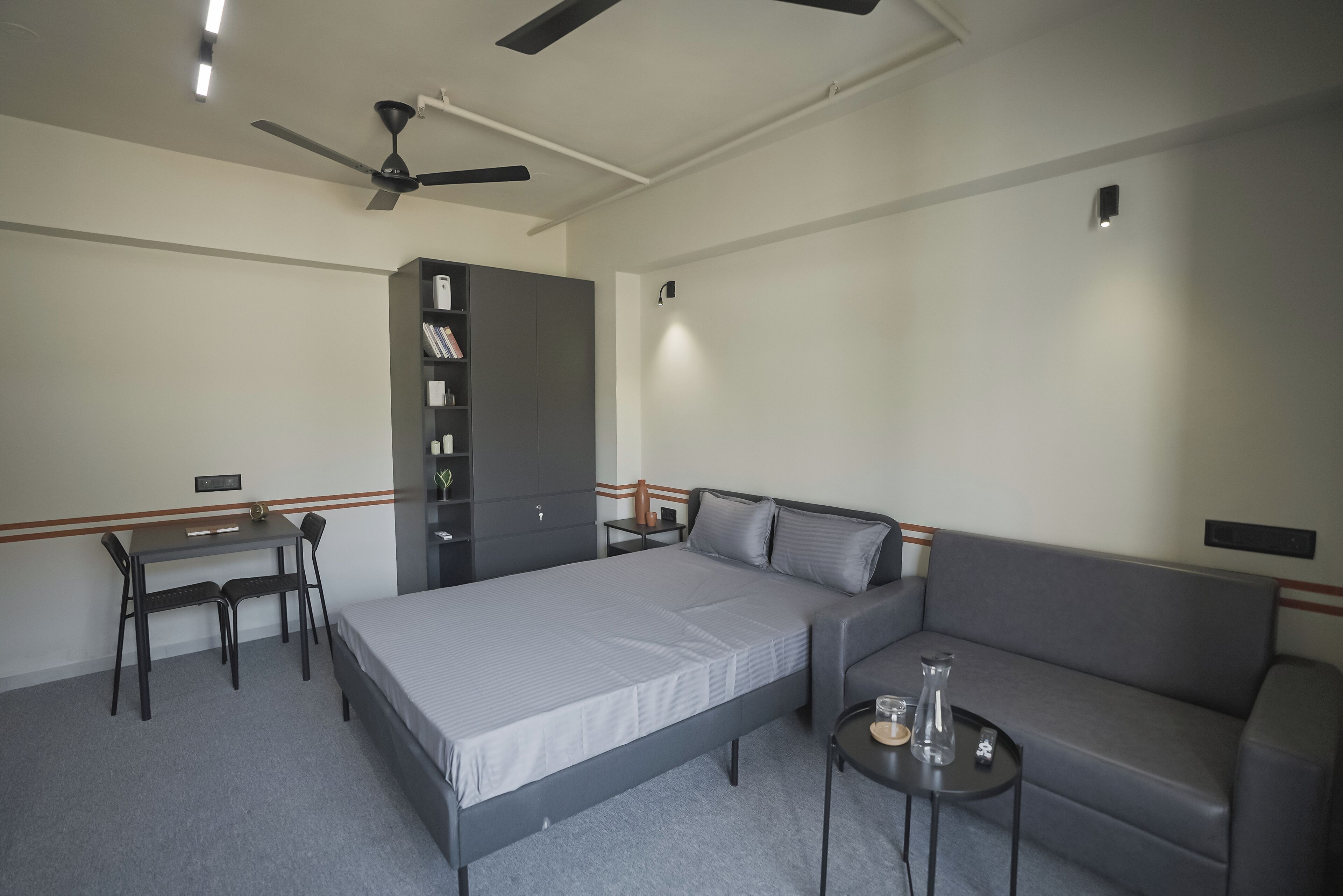 A bedroom with a bed, a desk, a chair, a sofa, a small table, a ceiling fan, and a wardrobe.
