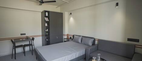 A bedroom with a bed, a desk, a chair, a sofa, a small table, a ceiling fan, and a wardrobe.