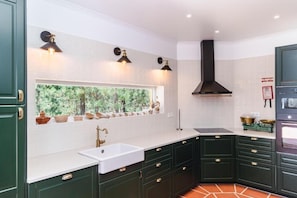 Una cocina con gabinetes verde oscuro, encimeras blancas, una ventana con vistas a la vegetación y una campana extractora negra.