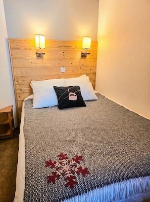 A bed with a gray blanket and a red snowflake design, two white pillows, a black pillow with a ski icon, wooden headboard, and two wall-mounted lights. - Thémis 112 - The Alpine Cocoon (La Plagne Tarentaise)