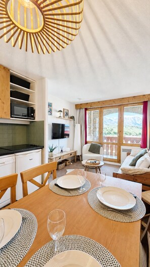 A dining area with a wooden table set for four, a television, and a view of a mountainous landscape through large windows. - Thémis 112 - The Alpine Cocoon (La Plagne Tarentaise)