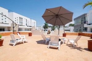 Eine Dachterrasse mit weißen Liegestühlen, einem großen Sonnenschirm und Blick auf moderne Gebäude.