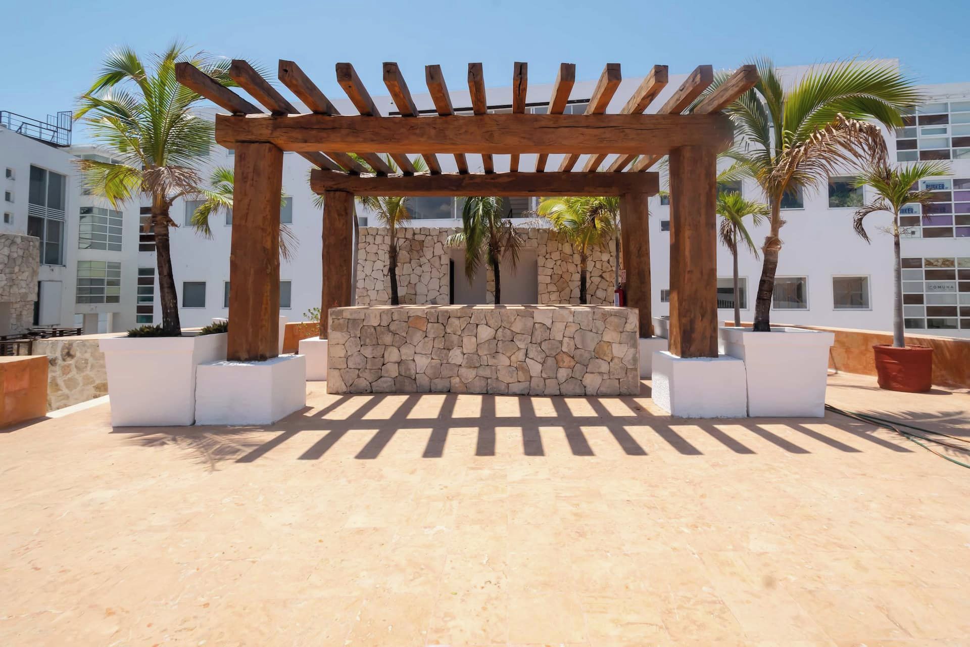 Une structure de pergola en bois, avec un mur de pierre au centre, entourée de plantes en pot et de palmiers, le tout se dressant sur fond de bâtiments modernes.