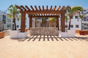 A wooden pergola structure with a stone wall in the center, surrounded by potted plants and palm trees, set against a backdrop of modern buildings. - Playa retreat w/ pool - walk to ruins, beach & 5th (Playa del Carmen)