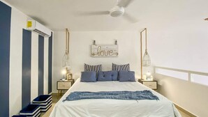 Quarto com cama, almofadas listradas, colcha branca, ventoinha no teto e um letreiro fixado na parede.