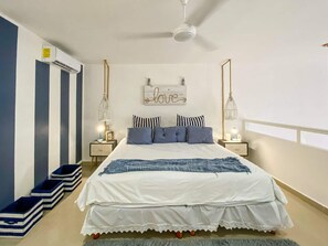 Ein Schlafzimmer mit einem Bett, gestreifte Kissen, einer weißen Tagesdecke, einem Deckenventilator und einem an der Wand befestigten Schild.