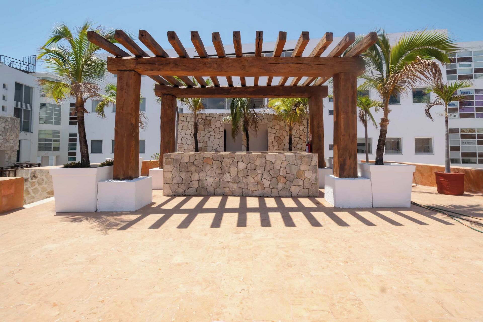 A wooden pergola structure with a stone wall base, surrounded by potted palm trees, set against a backdrop of modern buildings.