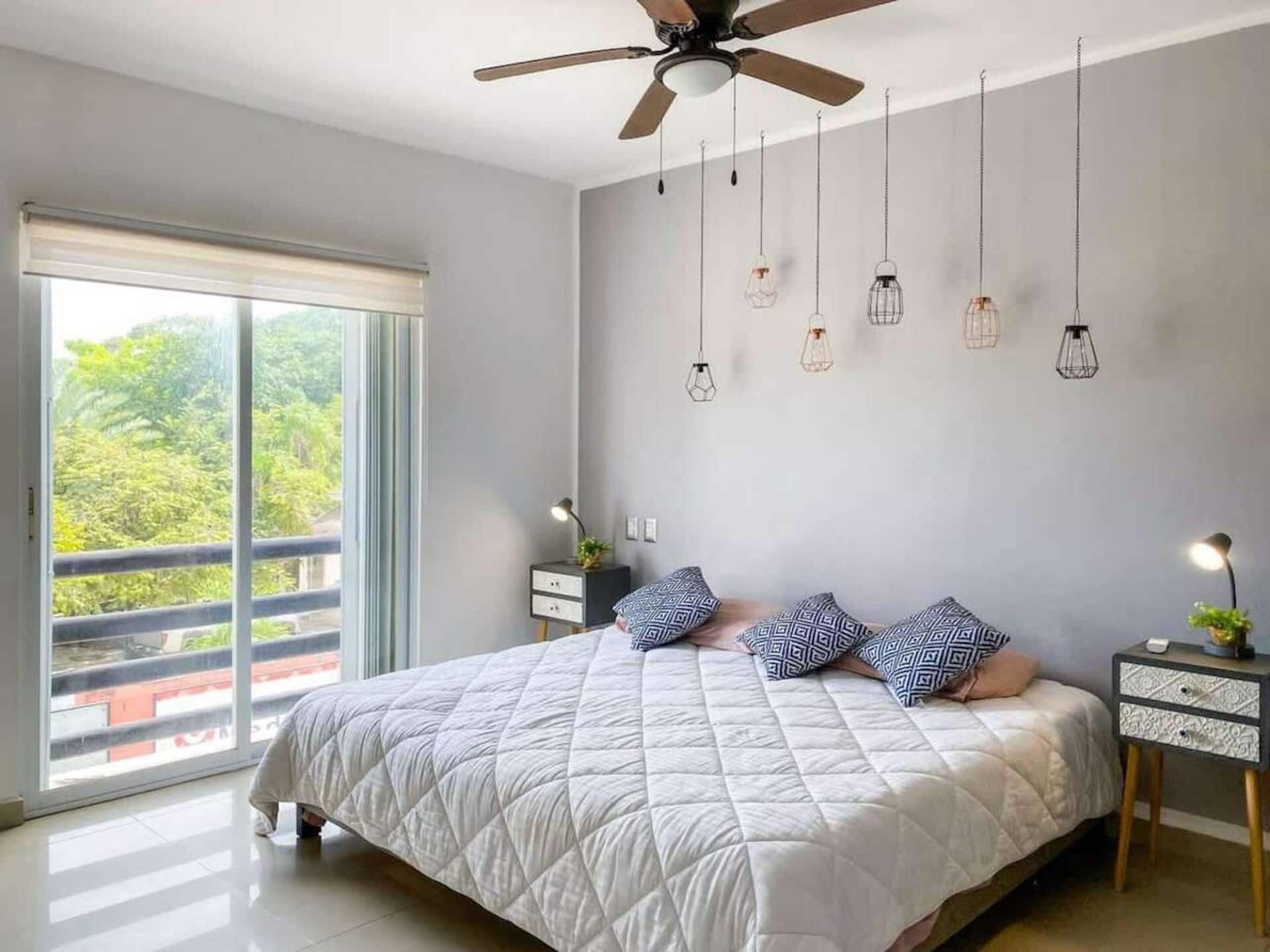 A bedroom with a bed, a ceiling fan, hanging light fixtures, a nightstand, and a view through a sliding door.