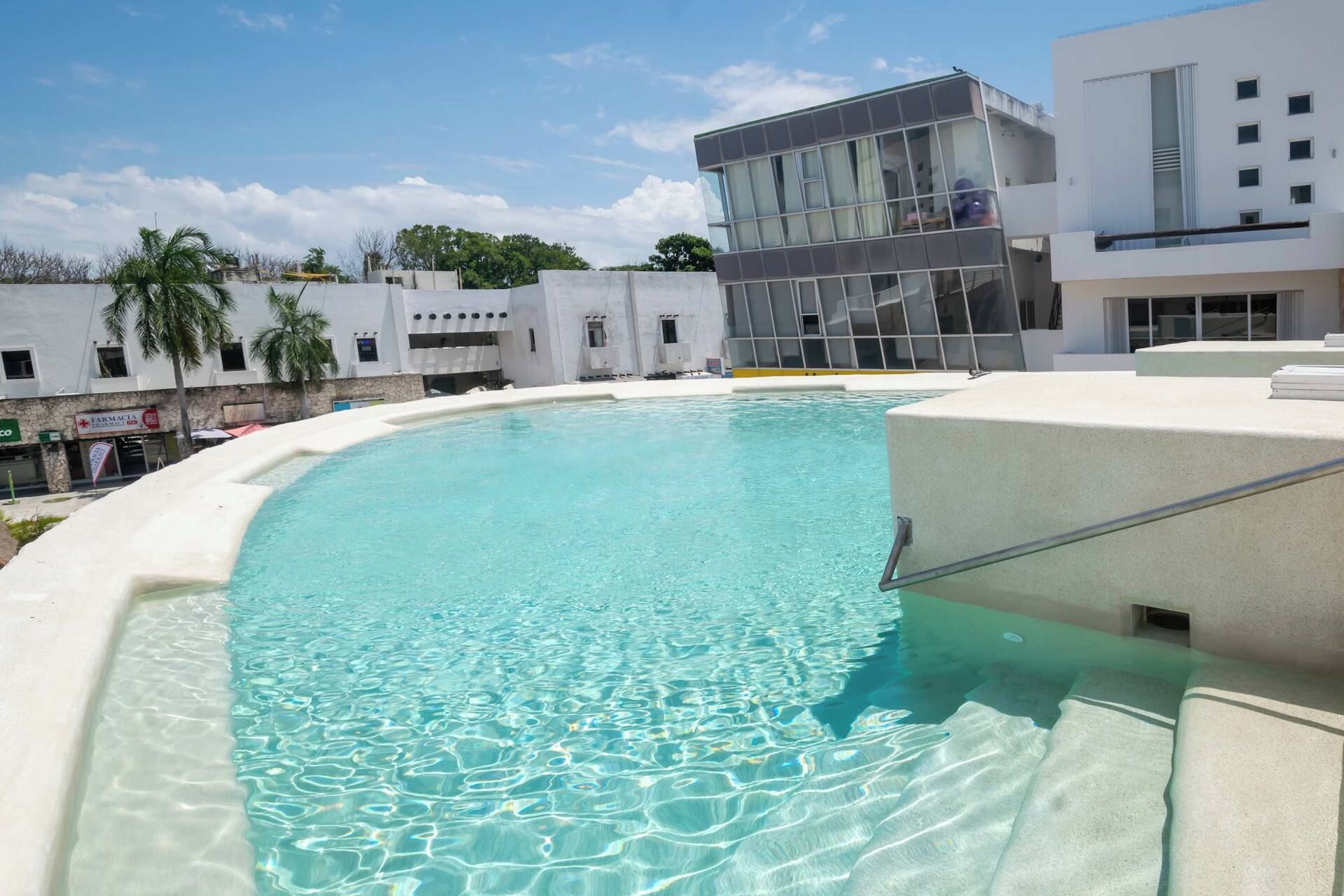 Ein klares Schwimmbecken mit gewölbter Kante, umgeben von einem weißen Gebäude mit modernem Design, und im Hintergrund ist eine Apotheke erkennbar.