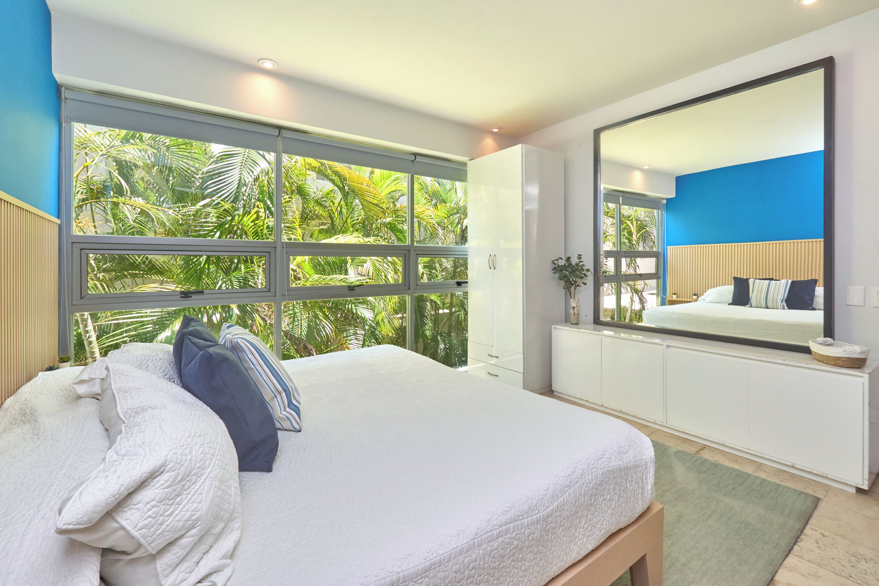 A bedroom with a large bed, a window with a view of greenery, a mirror, and a bedside table.