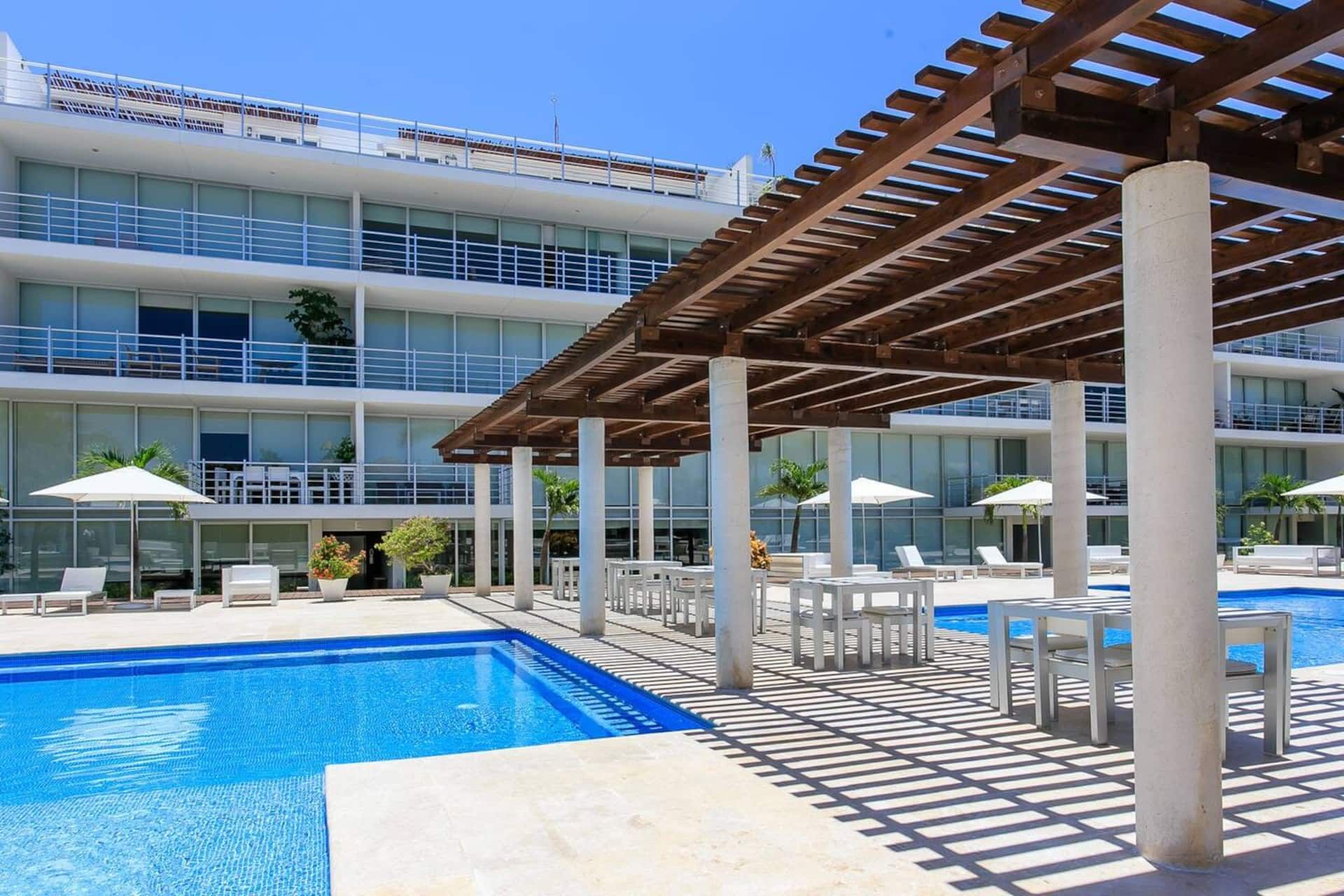 Un espace piscine doté de chaises longues et de parasols, un coin salon abrité et un bâtiment moderne à plusieurs étages avec des balcons.