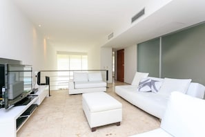 A modern living room with a flat-screen TV, white sofas, and a large window providing natural light. - Magia resort condo w/ pool & balcony (Playa del Carmen)