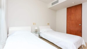 Hotellihuone, jossa on kaksi erillistä sänkyä, yöpöytä lampun kanssa, puinen ovi ja valkoinen seinä.