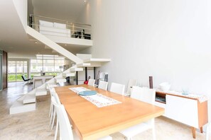 A spacious dining area with a long wooden table and white chairs, a staircase leading to an upper level, and a view of greenery outside. - Magia resort condo w/ pool & balcony (Playa del Carmen)