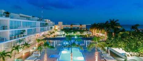 Un espace piscine d’hôtel aménagé sur plusieurs niveaux, agrémenté de palmiers et offrant une vue sur l’océan au crépuscule.