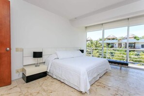 Ein modernes Schlafzimmer mit einem großen Bett, einem Nachttisch, einer Lampe und Blick auf eine tropische Landschaft durch große Fenster.