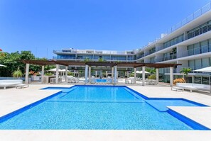 Ein Schwimmbecken mit einer Pergola, Liegestühlen und einem modernen Gebäude mit Balkonen im Hintergrund.