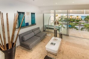 A modern living room with a gray sofa, a white coffee table, and a large window offering a view of a residential area. - Beachside penthouse w/ pool & hot tub (Playa del Carmen)