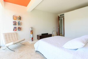 Ein Schlafzimmer mit einem Bett, einem Sessel, einem Nachttisch und einer Wand mit Kunstwerken.