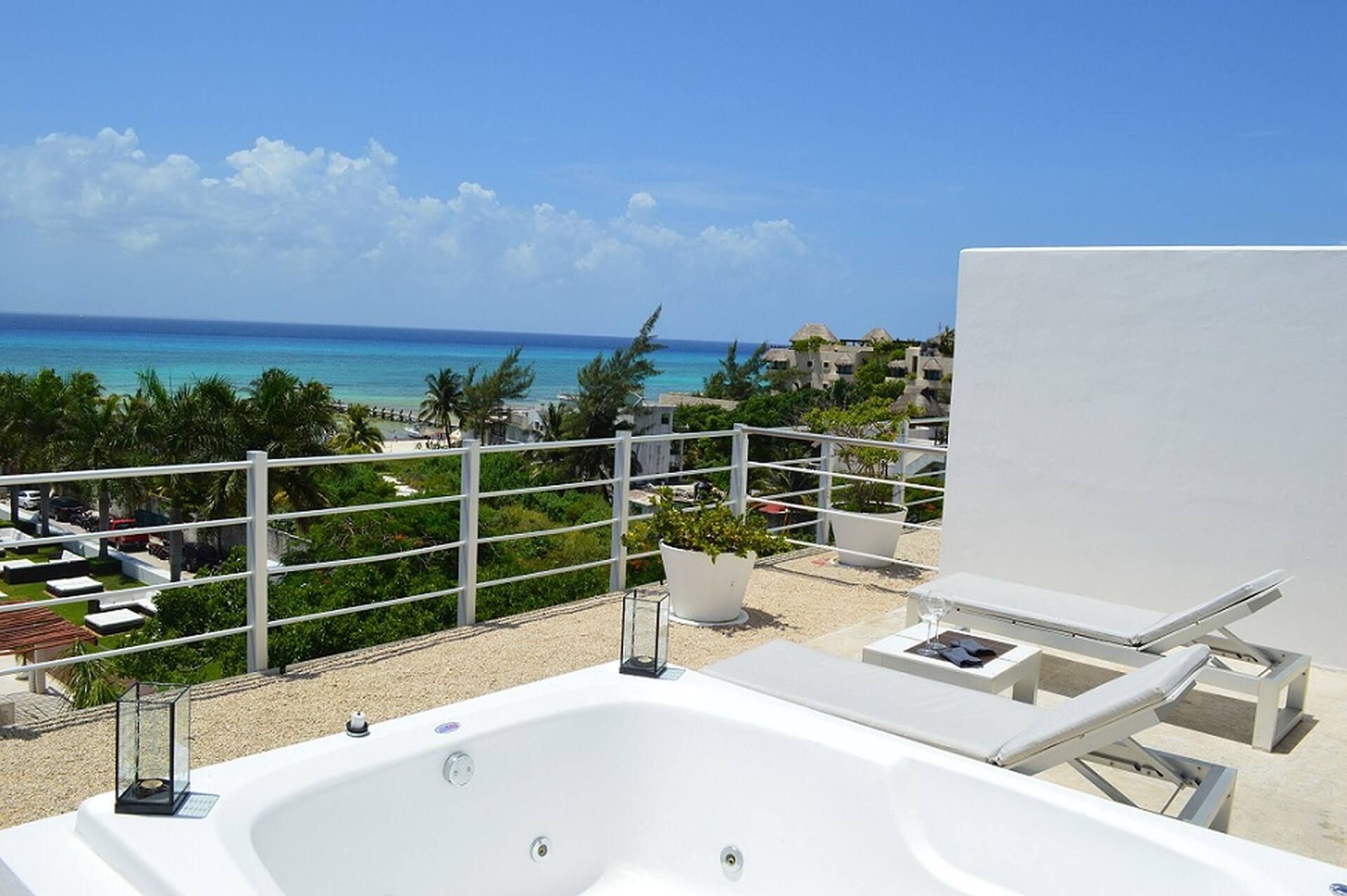 Un balcon avec une baignoire blanche, des chaises longues et une vue sur l’océan et les bâtiments.