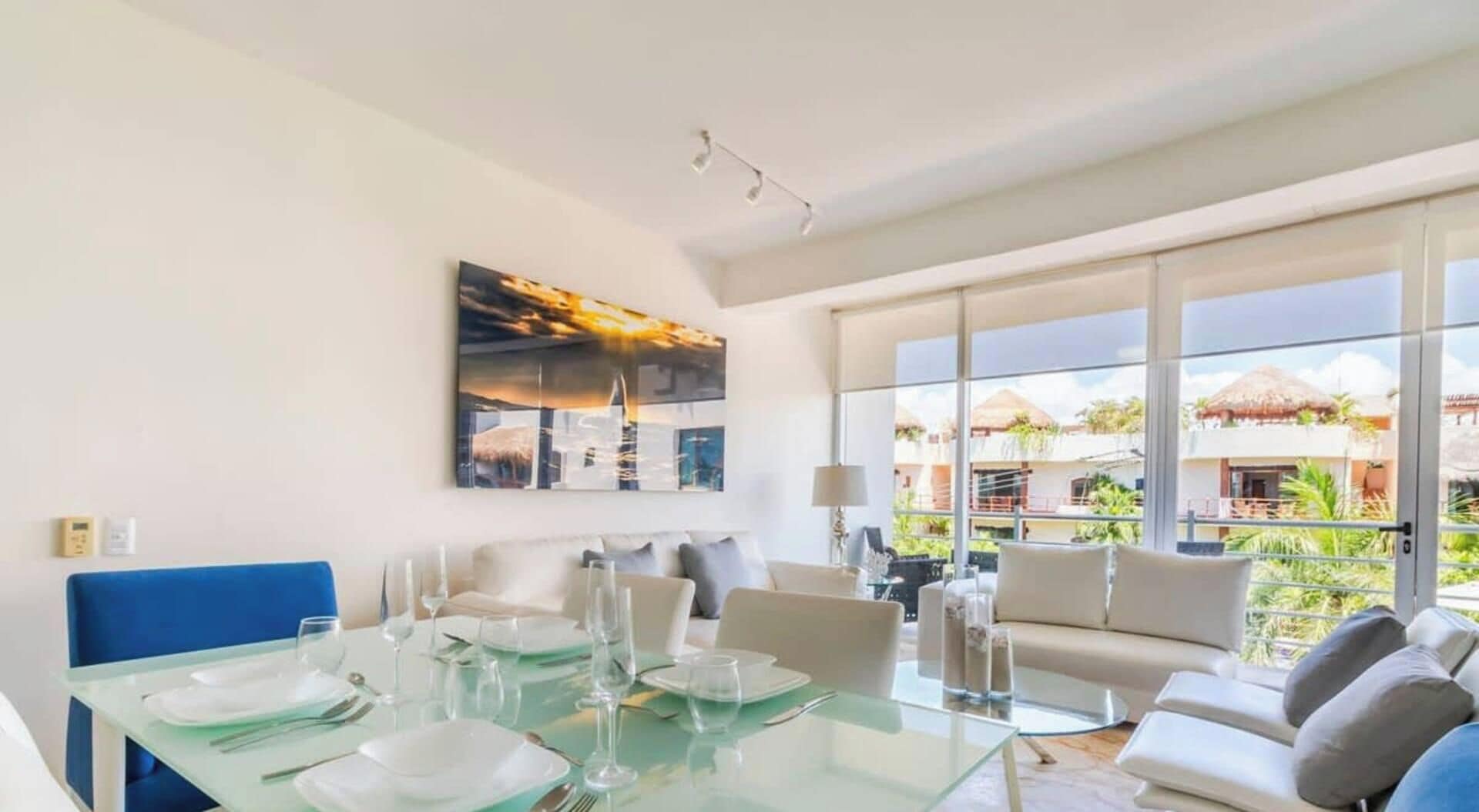 Ein moderner Essbereich mit einem Glastisch, der für eine Mahlzeit gedeckt ist, einer Couch und einem großen Fenster mit Blick auf eine tropische Landschaft.