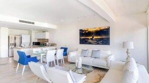 Ein modernes Wohnzimmer mit einer Couch, Sesseln, einem Glastisch und einem Essbereich mit einer Küche im Hintergrund.