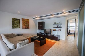 Ein modernes Wohnzimmer mit Sofa, Couchtisch, Fernseher, Kunstwerken und Blick ins Schlafzimmer.