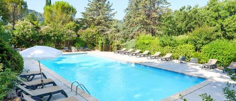 Una piscina rettangolare con superficie dell'acqua di un blu cristallino, circondata da una terrazza pavimentata. Sono presenti sedie a sdraio e una zona relax coperta con ombrelloni. La piscina è immersa in un giardino curato, con alberi e arbusti di varie specie.
