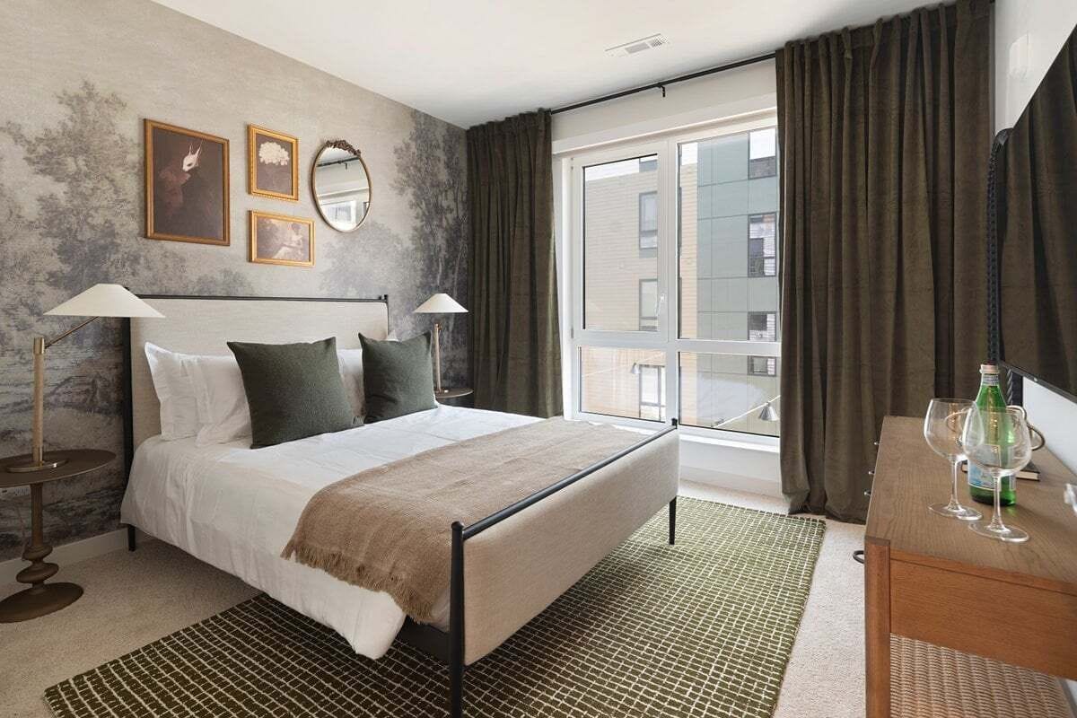 Habitación de hotel con cama, lámparas de noche, cómoda, ventana con cortinas y vistas al exterior.