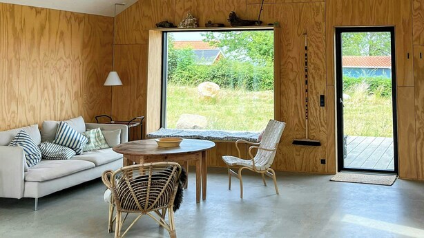 Et moderne interiør med skrå loft, trævægge, et stort vindue, en sofa, et spisebord og to fletstole.